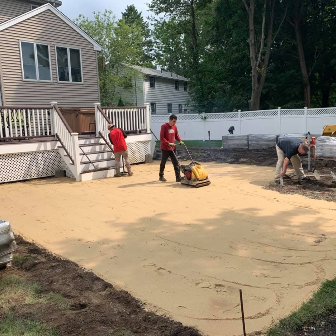 Crew compacting the sand base for a new paver patio behind a deck.