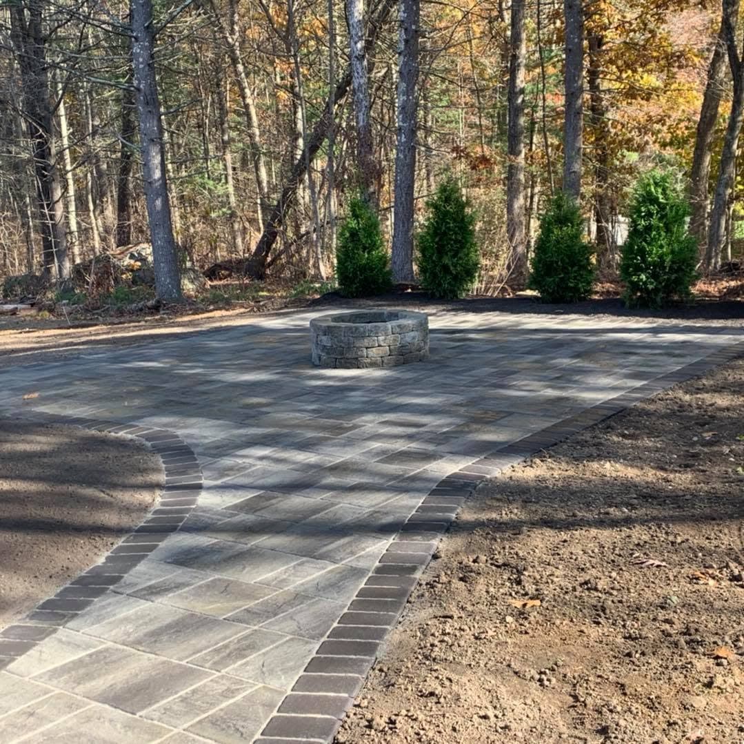 Paver patio with a round stone fire pit, reached by a curved paver walkway, with new arborvitae planted behind.