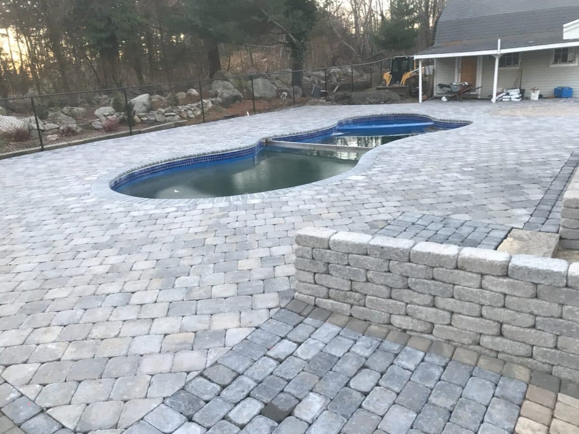 Fitted block retaining wall stepping down a yard beside a paver pool patio.