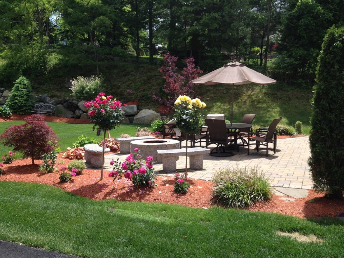 Paver patio with an umbrella and seating, edged by mulched beds of shrubs and flowering plantings beside a cut lawn.